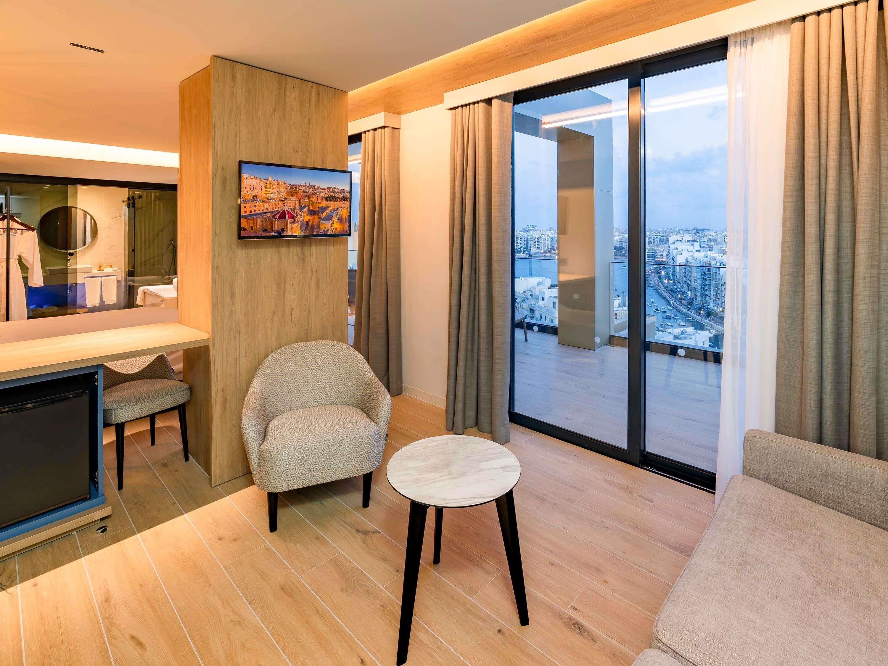 A modern hotel room with a city view, a round table, a sofa, and a small armchair.