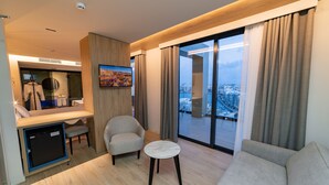 A modern hotel room with a sofa, armchair, small round table, and a view of the city through large windows. - Mercure St. Julian's Malta (St. Julian's)