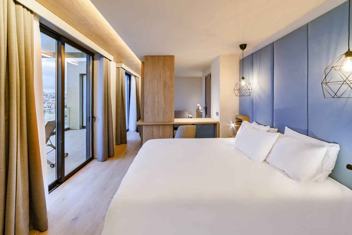 A modern hotel room with a large bed, a desk, and a view of the city through floor-to-ceiling windows.