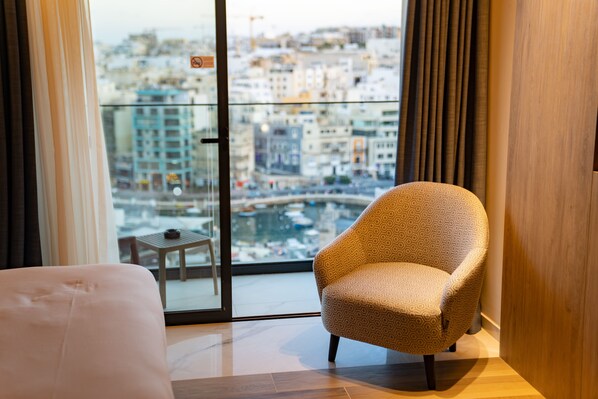 A hotel room with a large window offering a city view, a patterned armchair, and a small table with a cup. - Mercure St. Julian's Malta (St. Julian's)
