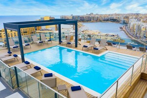 Una piscina infinita en la azotea con sillones de descanso y barandilla de cristal con vistas a la ciudad costera.