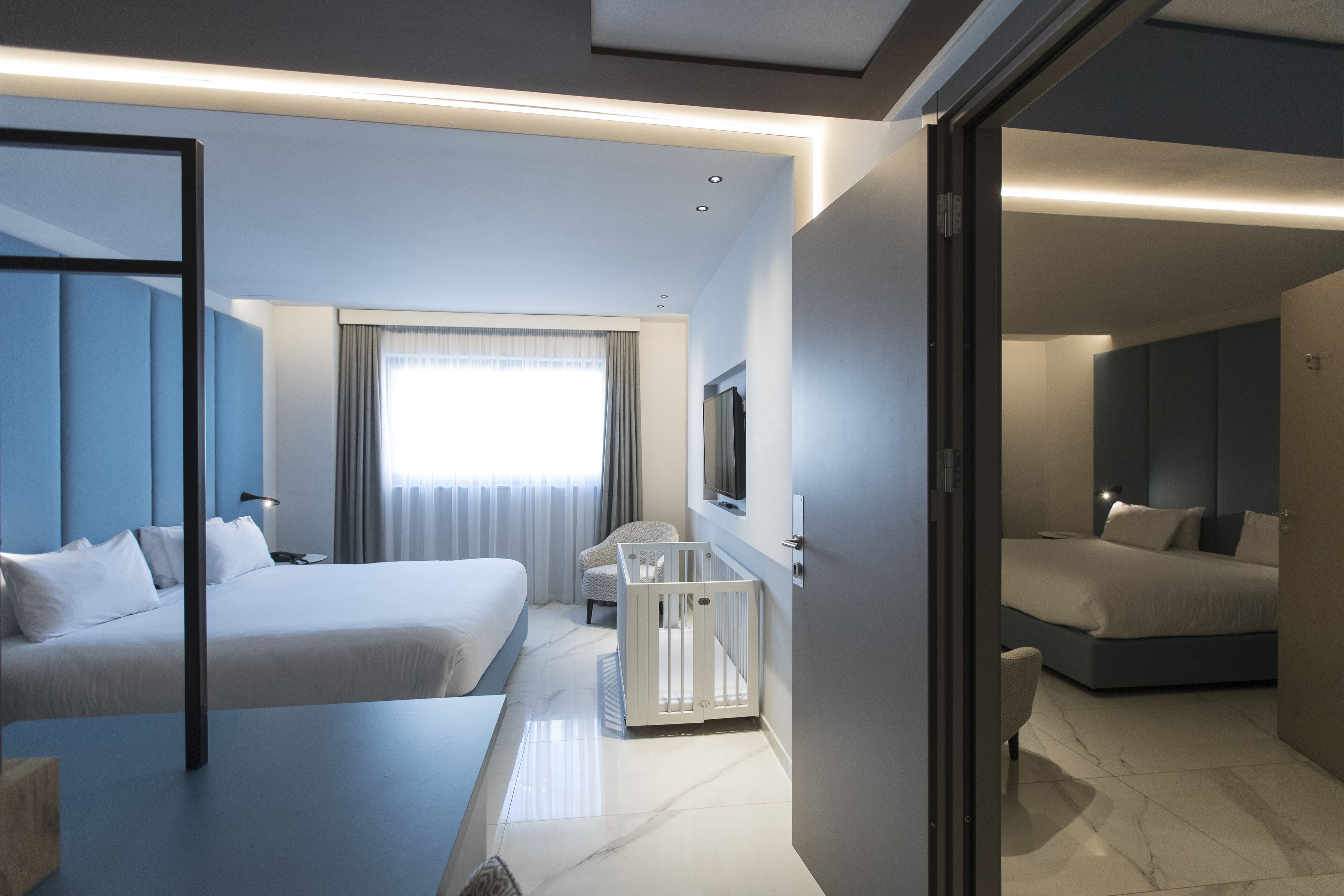 A modern hotel room with a large bed, a smaller bed, a desk, and a crib.