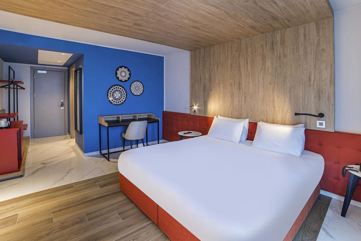 A modern hotel room with a large bed, a desk, and a red accent wall.