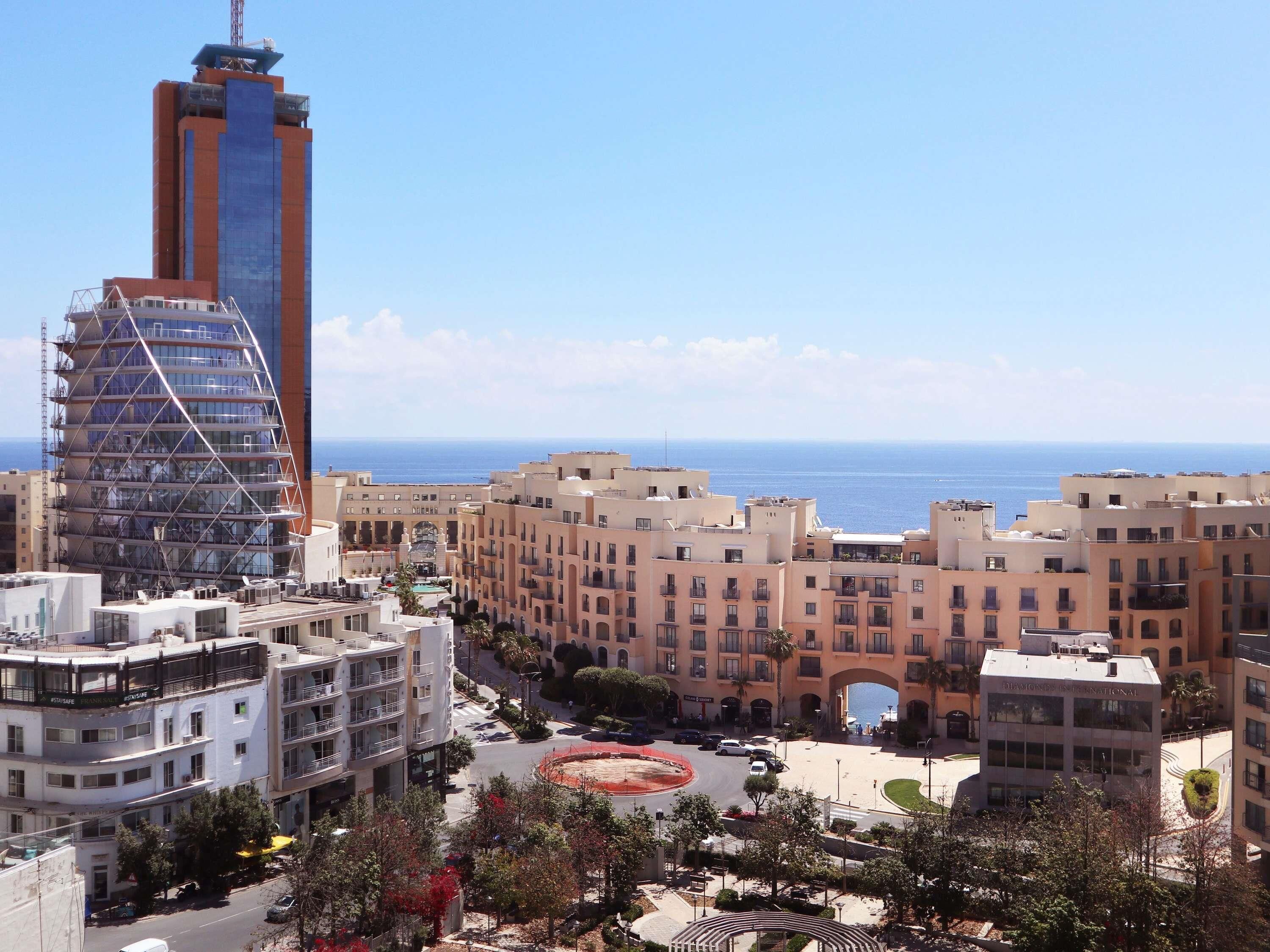 Un paysage urbain côtier avec des bâtiments modernes, un gratte-ciel imposant en construction et une vue dégagée sur la mer.