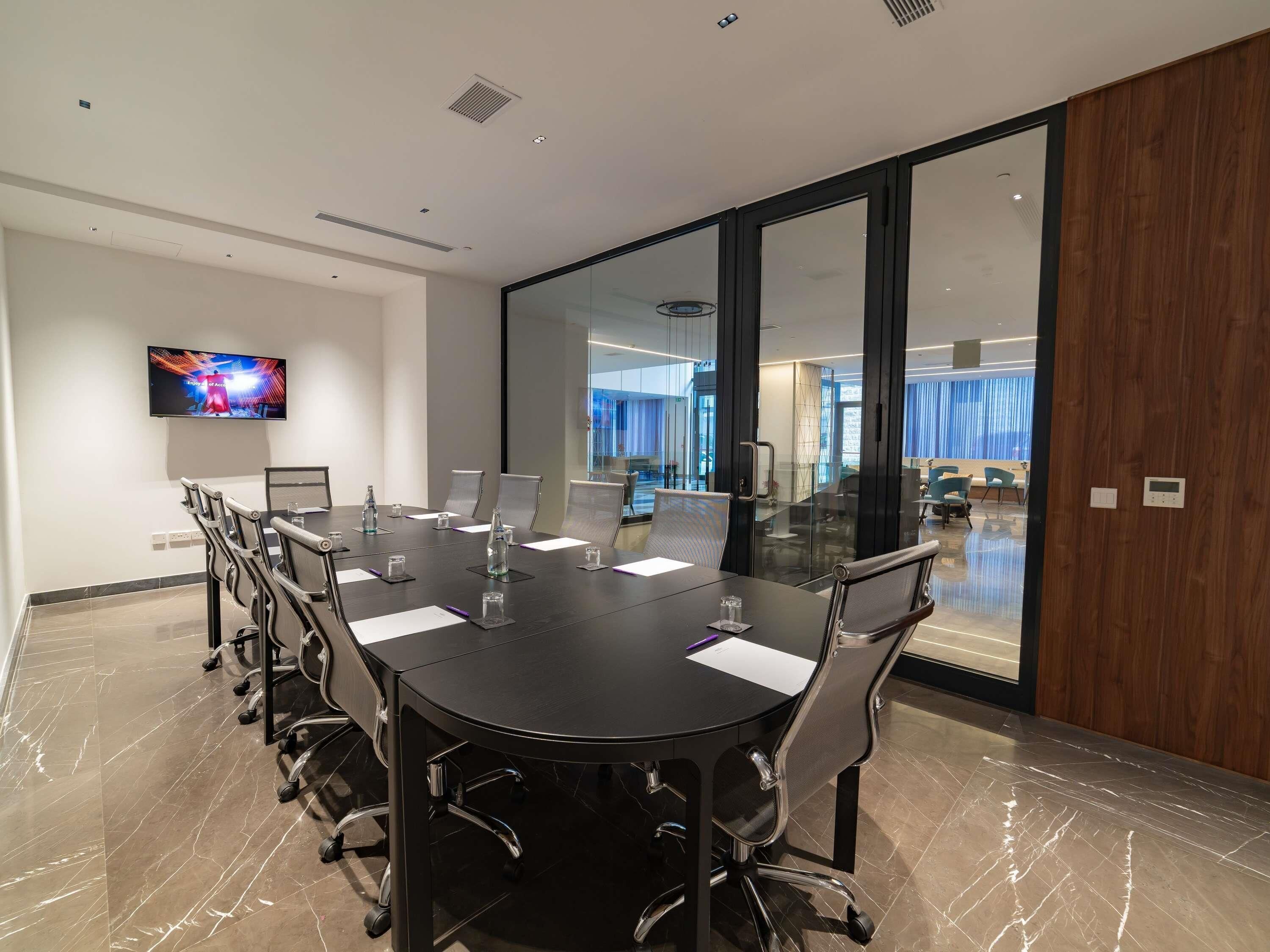 A conference room with a large table, chairs, a television, and a view of a modern interior space.