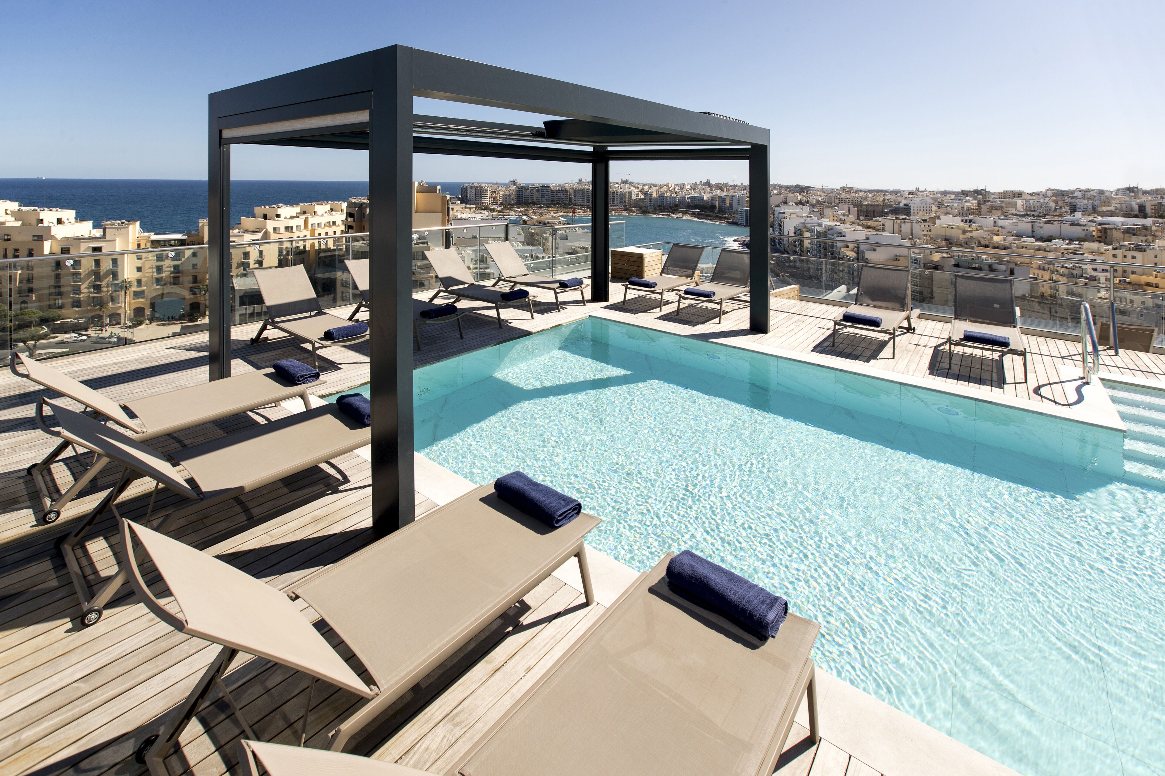 Una piscina sul tetto con poltrone relax e un recinto in vetro che si affaccia su una vista della città e del mare.