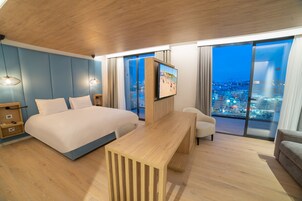 Una habitación de hotel moderna con una cama grande, un escritorio de madera, un televisor y vista a la ciudad desde la ventana.