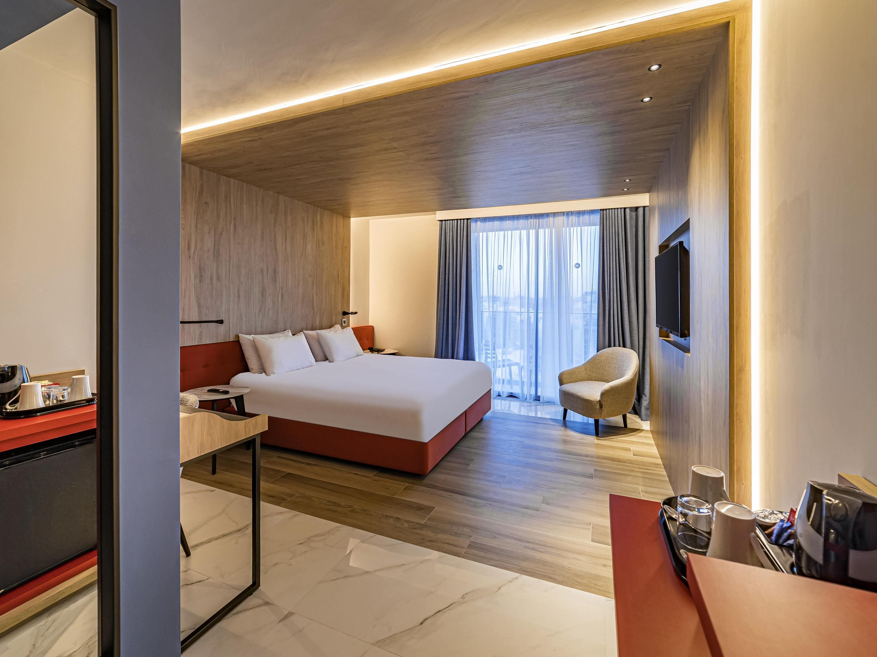 Habitación de hotel moderna con una cama grande, un escritorio con una silla, un televisor montado en la pared y vistas al exterior a través de cortinas translúcidas.