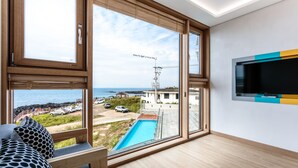 A modern room with large windows overlooking a pool and ocean, a flat-screen TV mounted on the wall, and a wooden bench with patterned cushions. - Jeju Ollero (Jeju City)