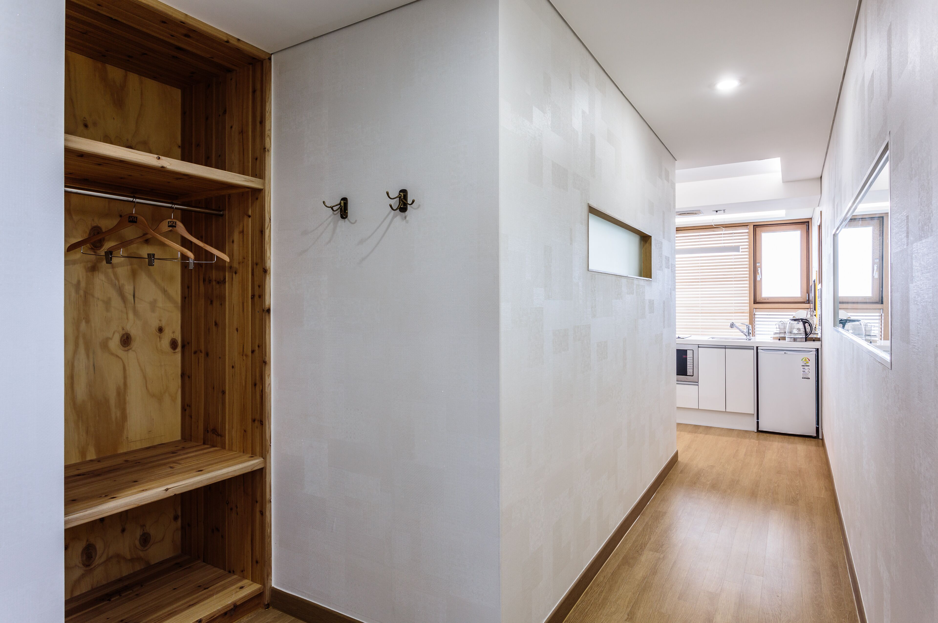 A hallway with a wooden wardrobe, a kitchen area with a sink and cabinets, and a window with blinds.