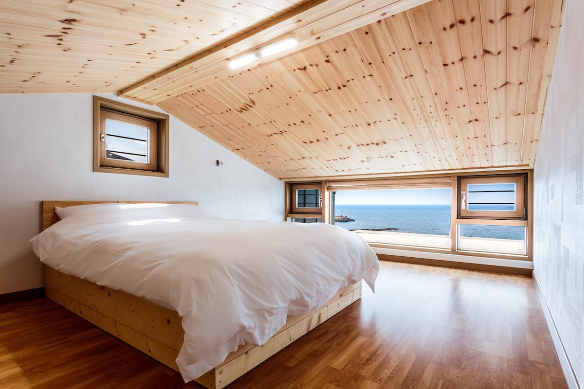 A bedroom with a wooden ceiling, a bed, and a view of the sea through large windows.