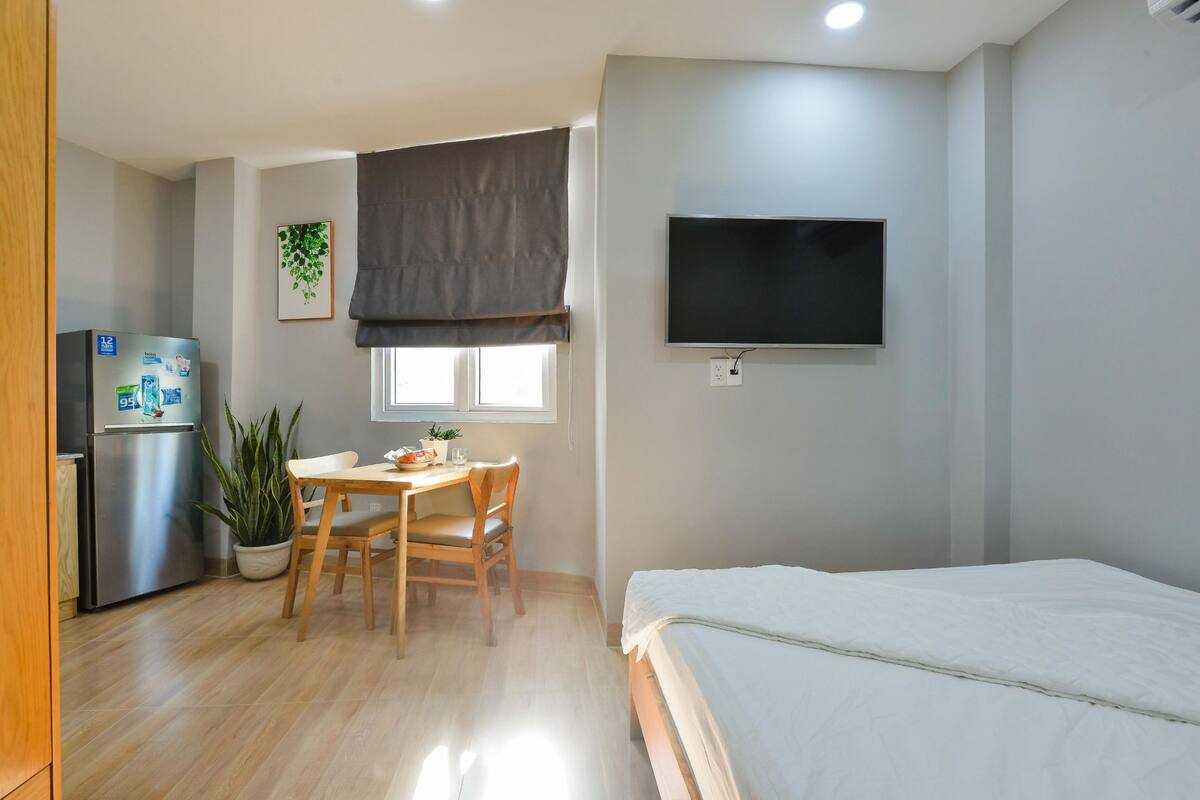 Een compacte kamer met een bed, een klein eettafeltje met stoelen, een koelkast, een potplant en een aan de muur bevestigde televisie.
