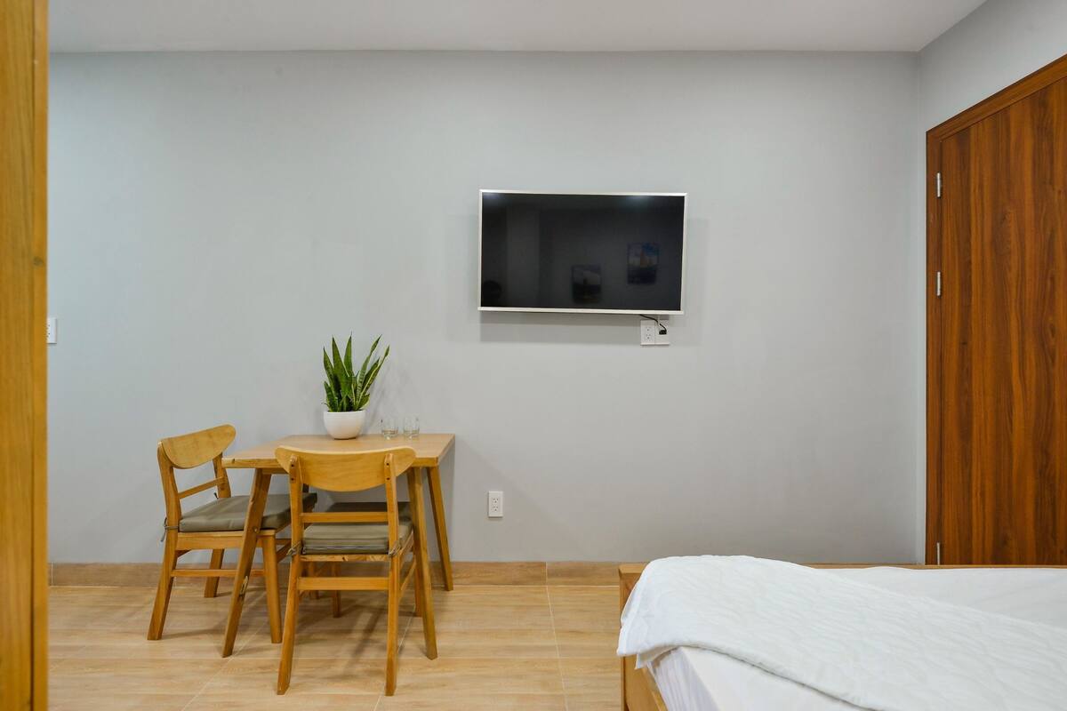 Een slaapkamer met een bed, een bureau met twee stoelen, een kamerplant en een wandtelevisie.