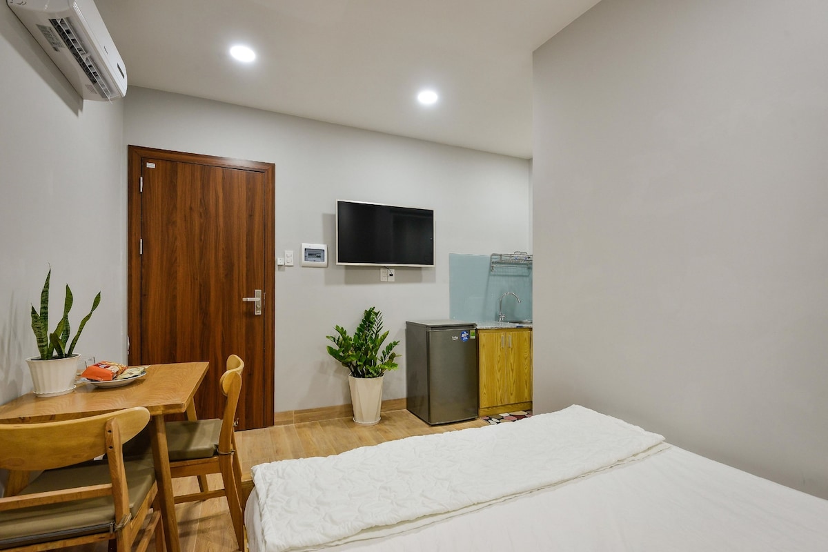 Een kleine kamer met een houten bureau, een stoel, een bed, een televisie, een koelkast en twee potplanten.