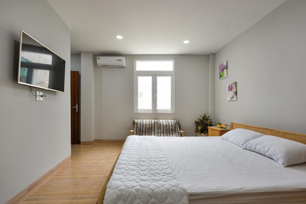 Een slaapkamer met een groot bed, een aan de muur bevestigde televisie, een raam en een klein nachtkastje met een plant.