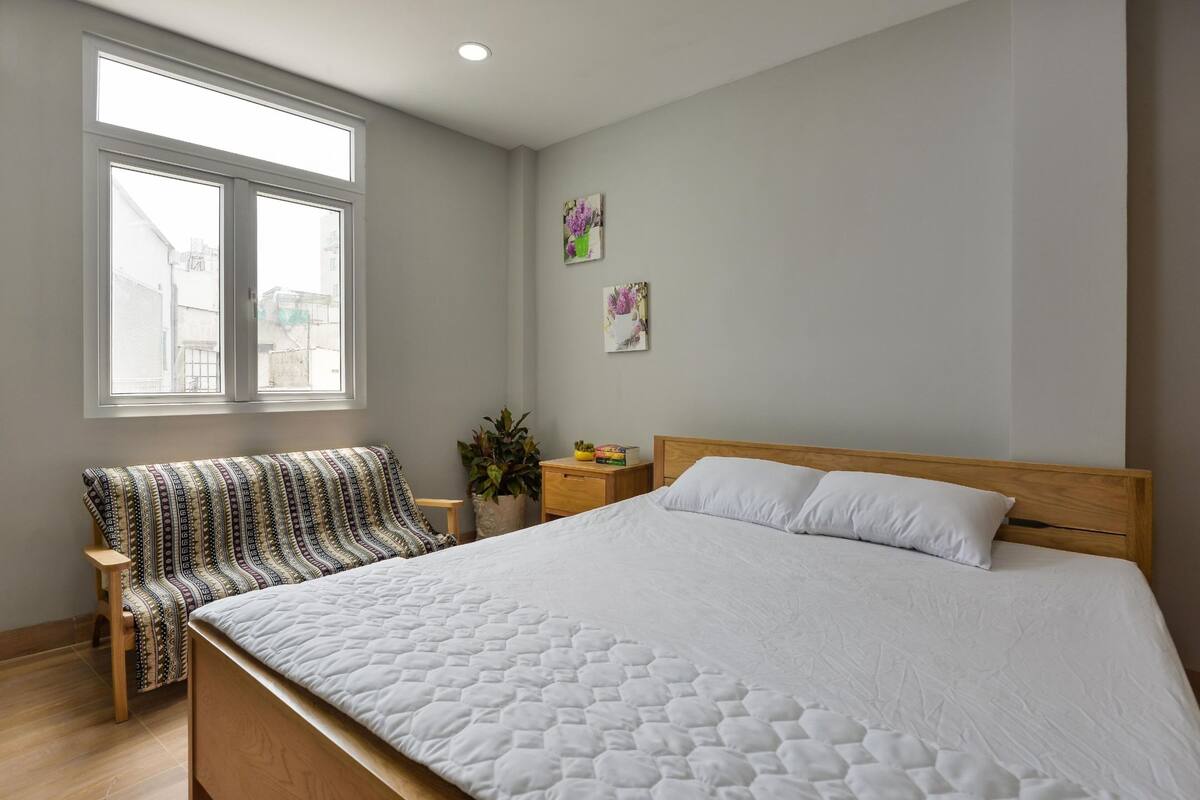 Een slaapkamer met een groot bed, een bankje, een nachtkastje en een raam met uitzicht op gebouwen.