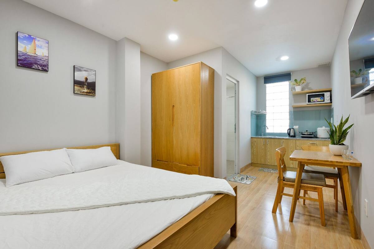 Een slaapkamer met een bed, een houten garderobe, een eettafel met stoelen en een keuken met een magnetron en een plant.