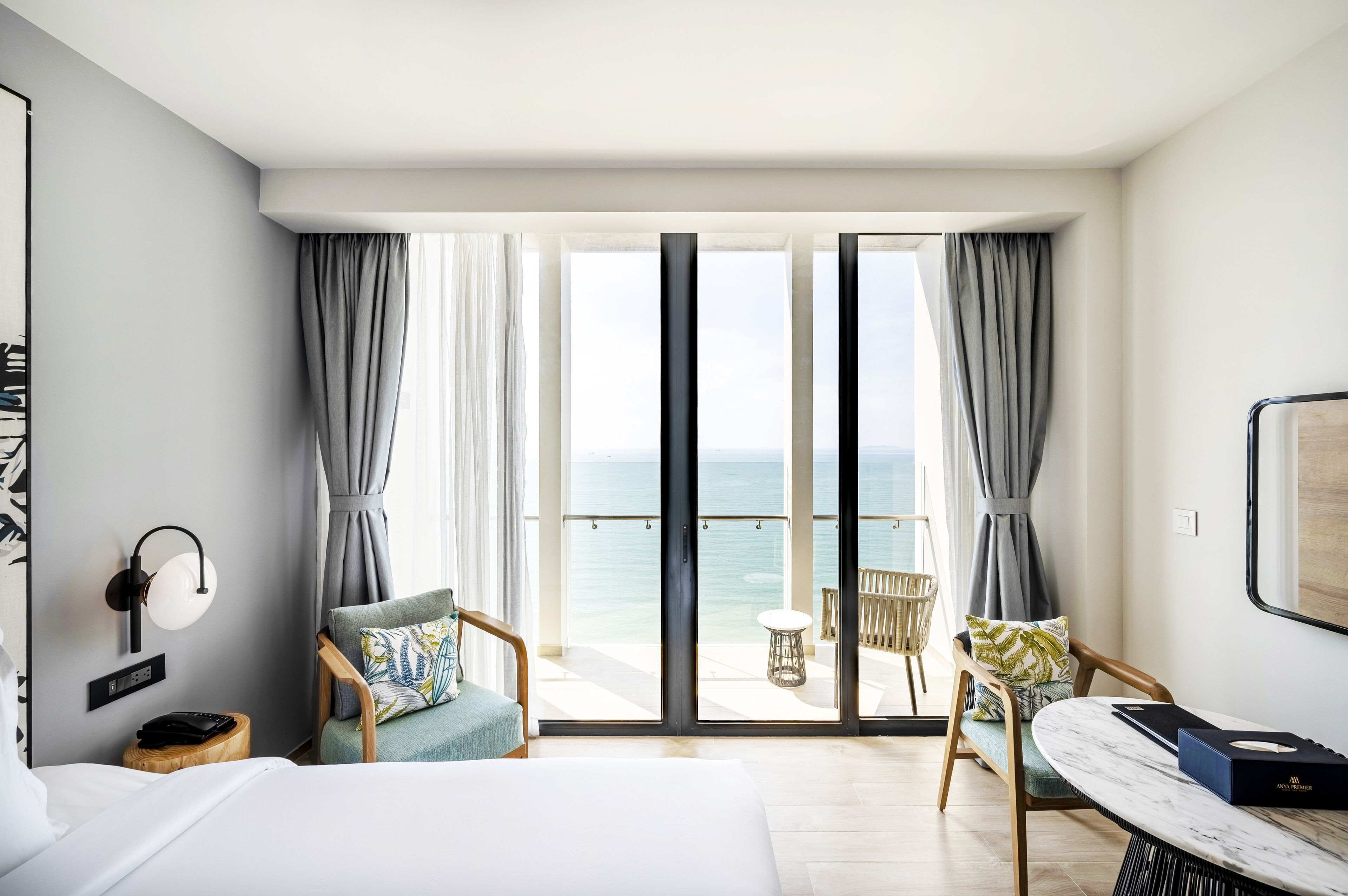 A modern hotel room with a large bed, a desk, and a view of the sea through floor-to-ceiling windows.