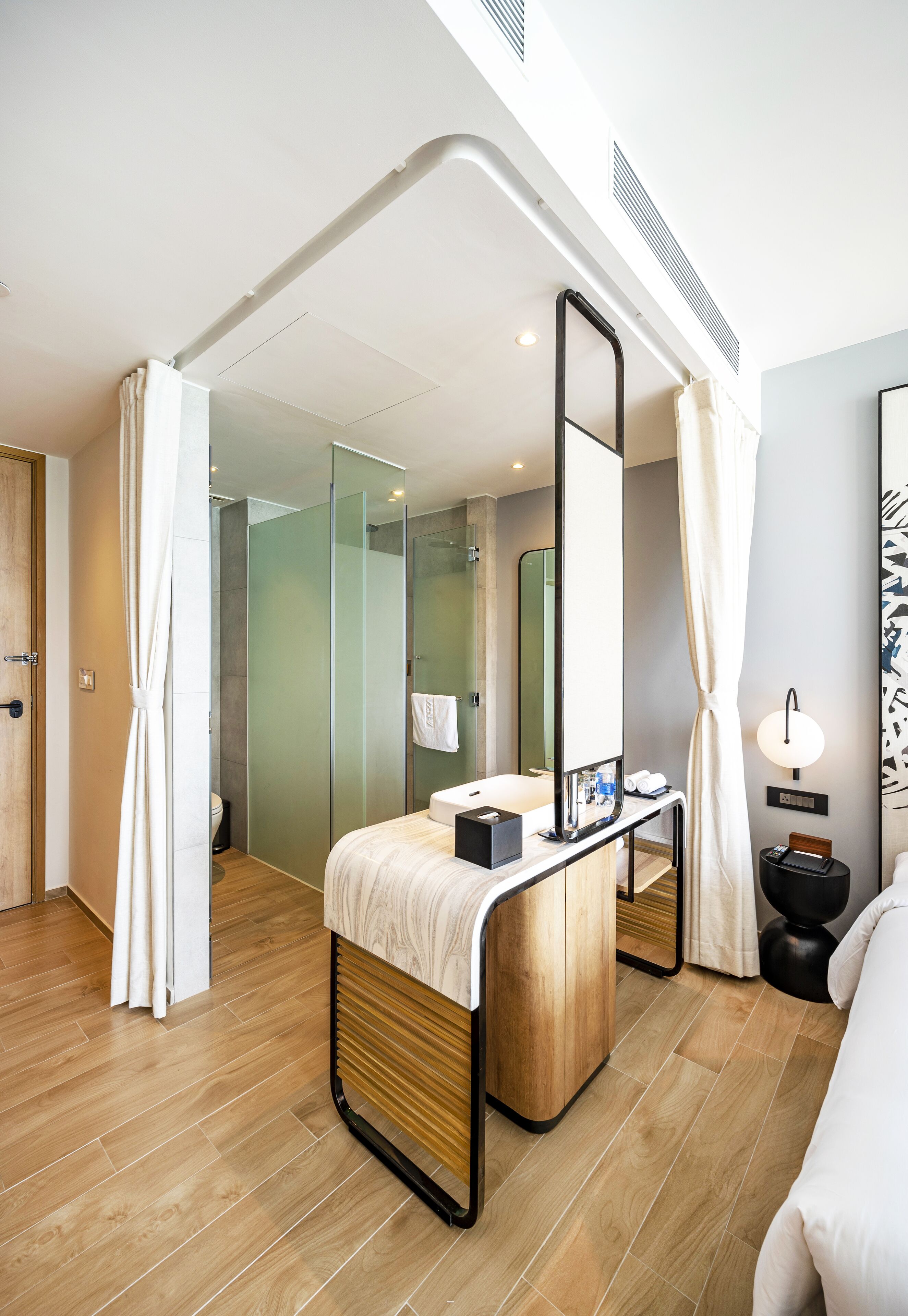 Ein modernes Hotelzimmer mit Holzboden, einem großen Bett, einem Nachttisch mit Lampe, einer Glasdusche und einem großen Spiegel.