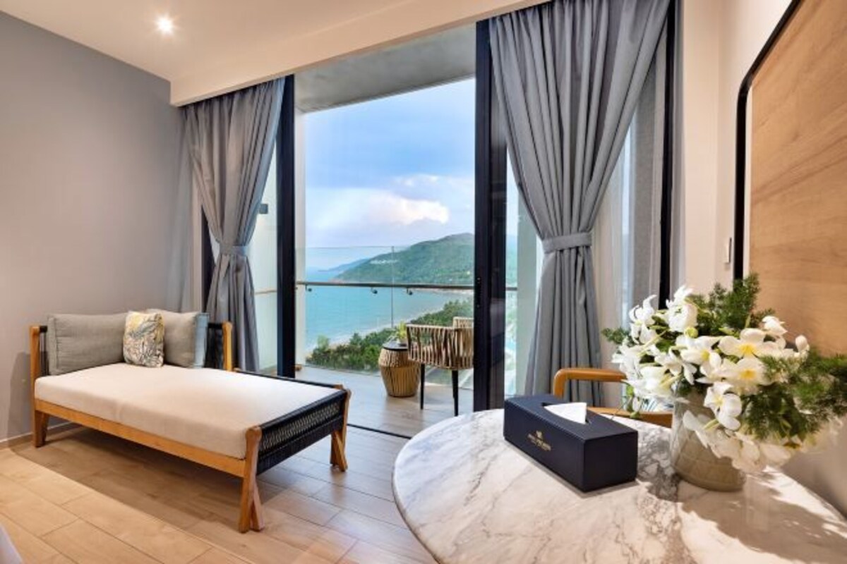 A bedroom with a bed, a marble side table, a vase of flowers, and a view of the outdoors.