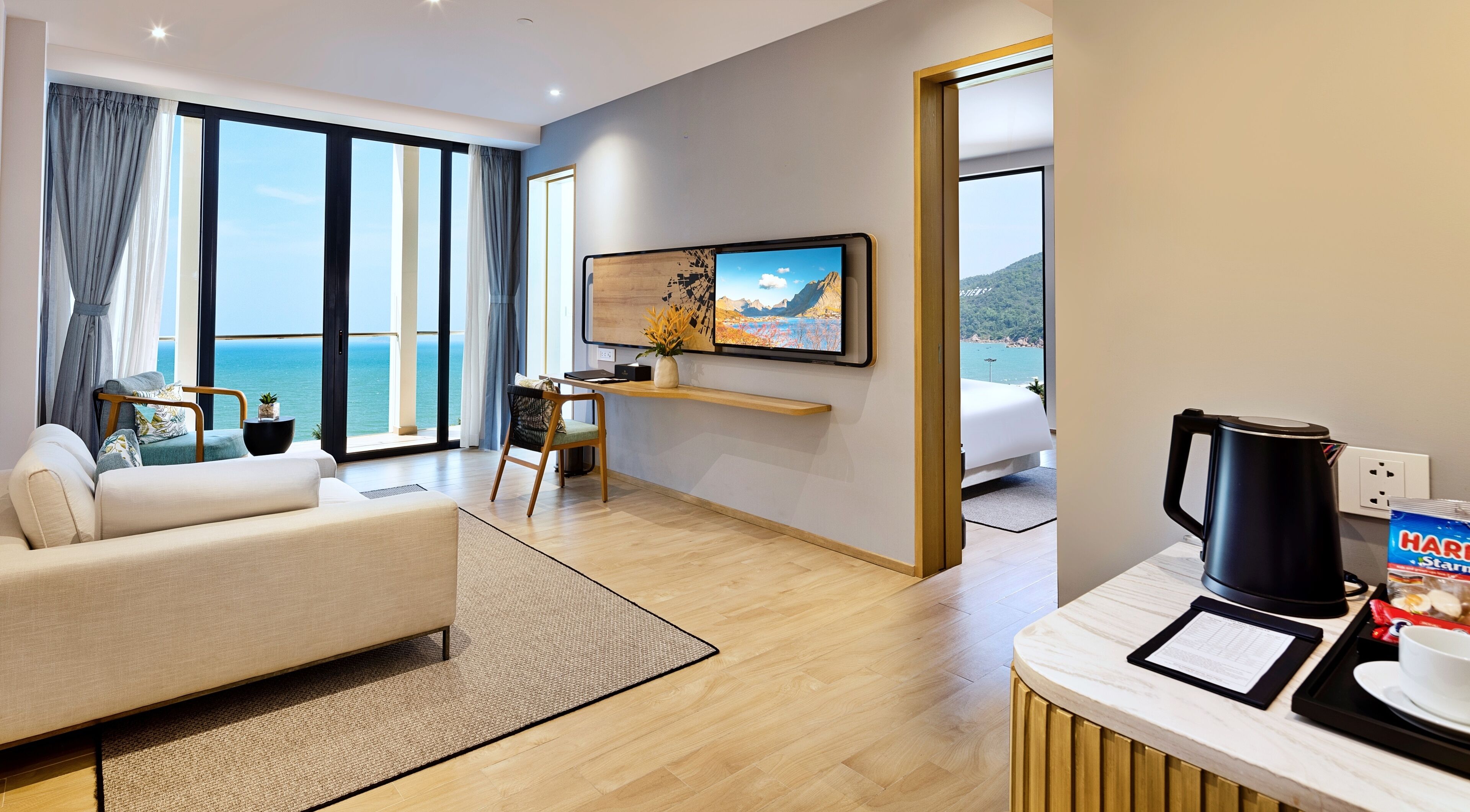 Ein modernes Hotelzimmer mit großem Fenster und Meerblick, einer bequemen Couch, einer kleinen Essecke und einer Küchenzeile mit Kaffeemaschine.