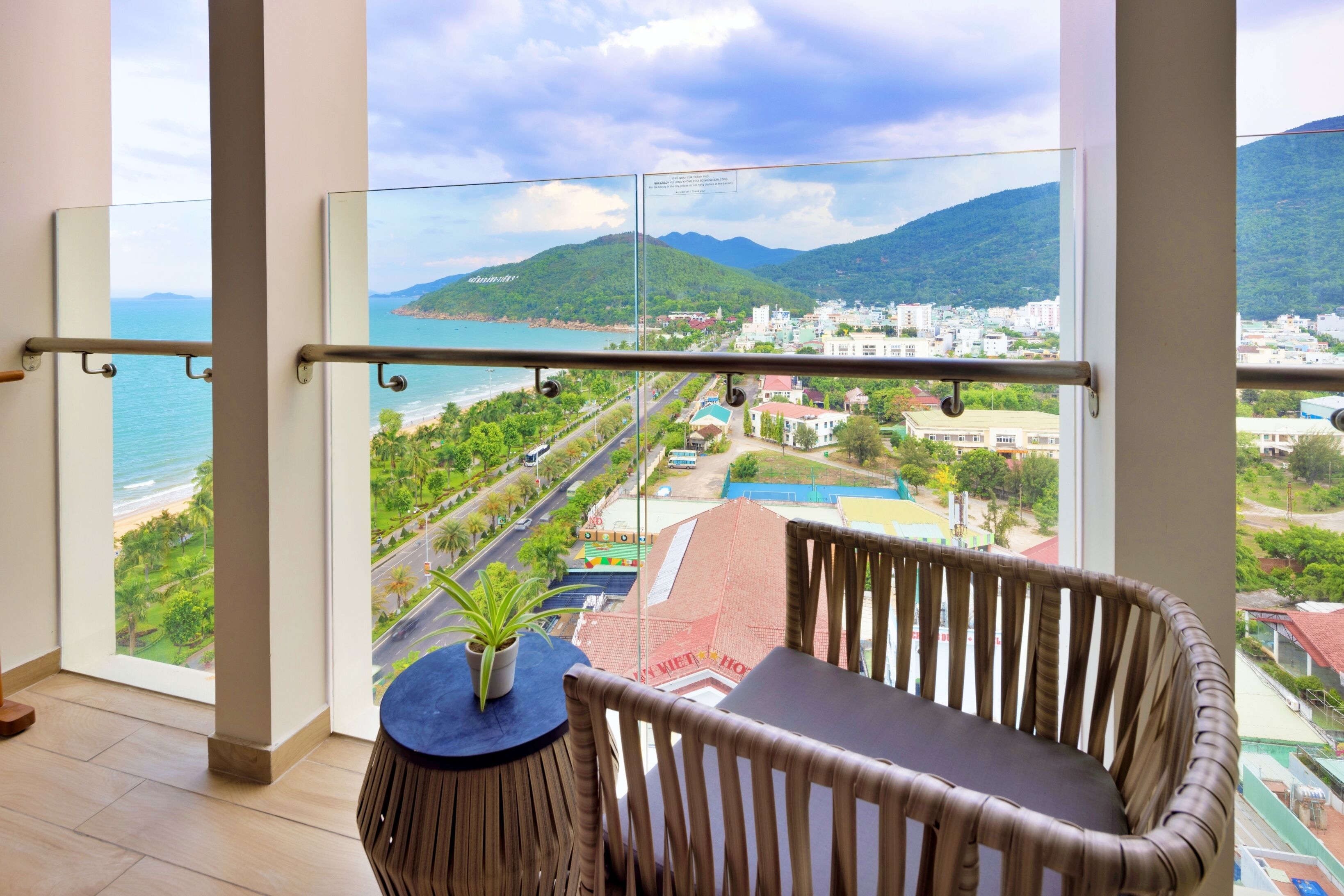 A balcony with a view of a beach and a cityscape, a round wooden table with a potted plant, and a wicker chair.