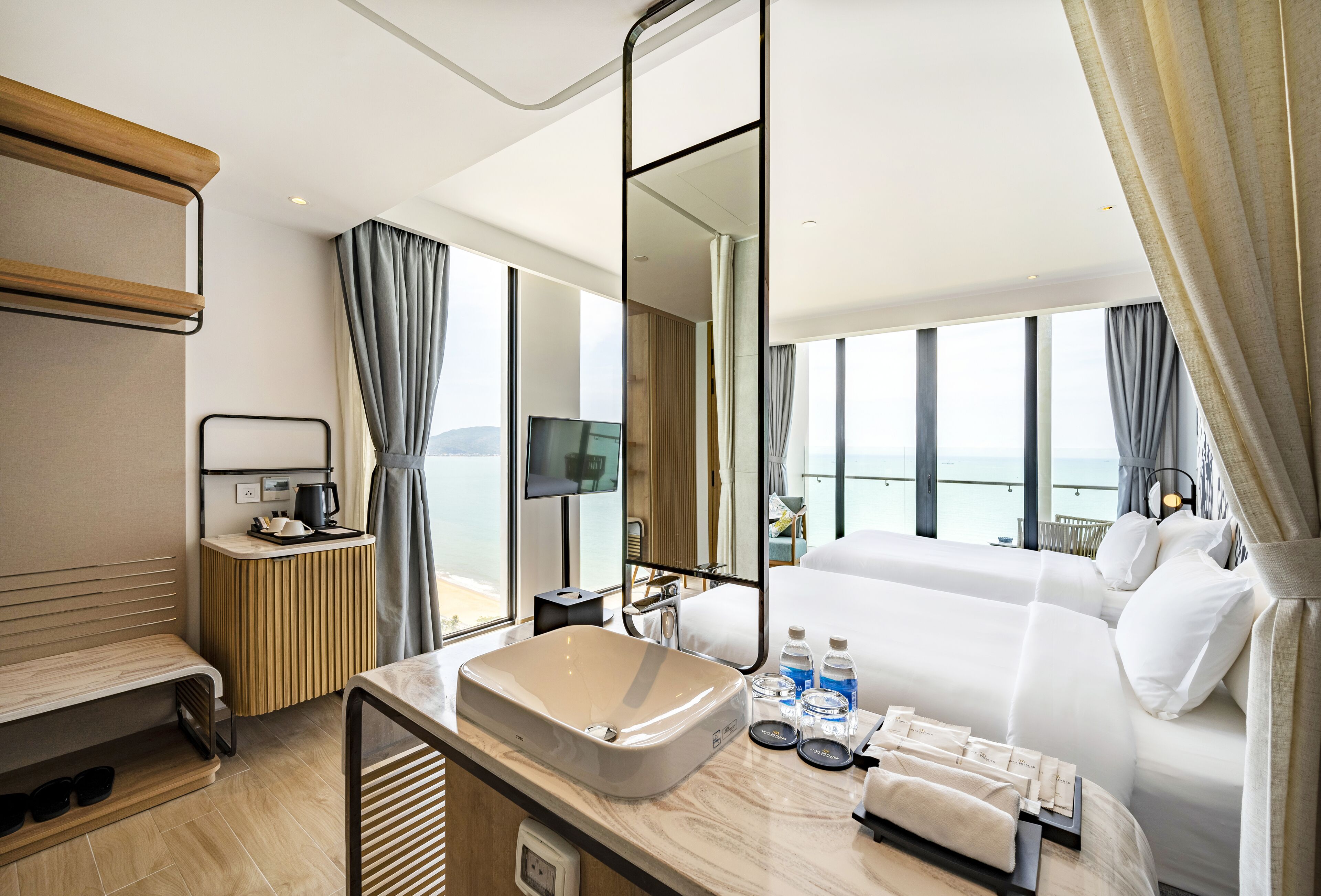 Ein modernes Hotelzimmer mit einem großen Bett, einem Badezimmer mit Waschbecken und Spiegel, einem Nachttisch mit Pflegeprodukten und Blick auf das Meer.