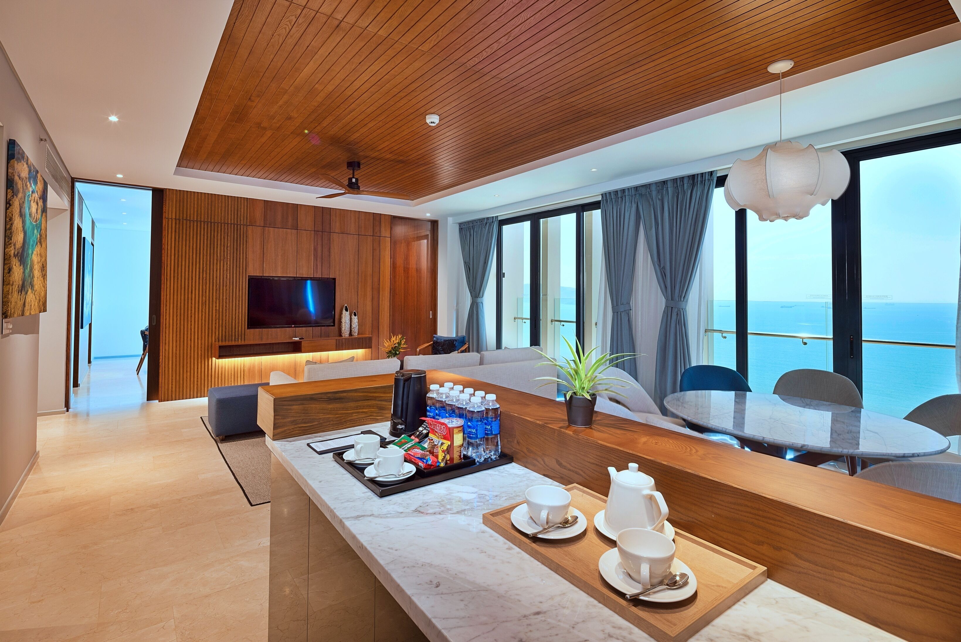 A modern hotel room with a wooden ceiling, a large TV, a dining area with a table and chairs, and a view of the sea.