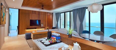 A modern hotel room with a wooden ceiling, a large TV, a dining area with a table and chairs, and a view of the sea.