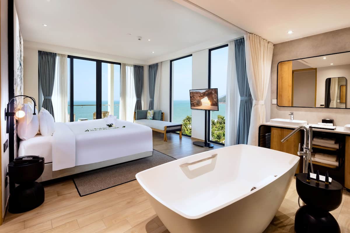 Ein modernes Hotelzimmer mit einem großen Bett, einer Badewanne, einem Balkon mit Meerblick und einem an der Wand befestigten Fernseher.