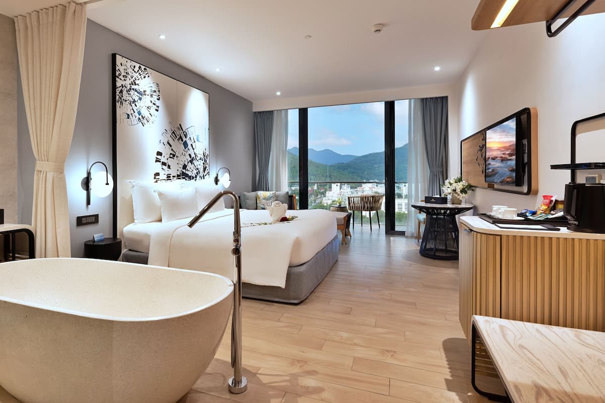 Ein modernes Hotelzimmer mit einer großen Badewanne, einem Bett mit weißen Leinentüchern, einem Balkon mit Blick auf die Stadt und einem Essbereich mit Tisch und Stühlen.
