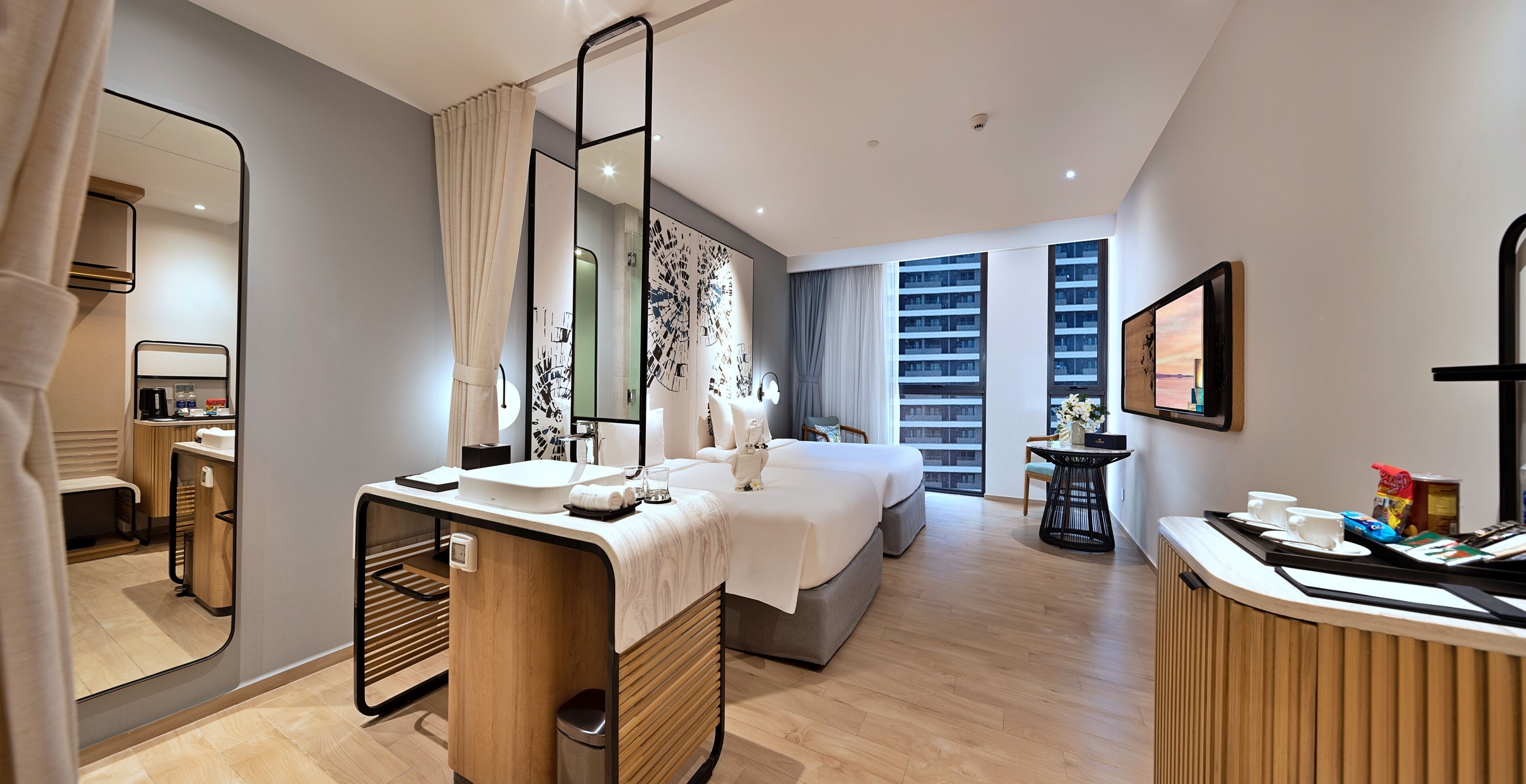 A modern hotel room with a large bed, a desk with a mirror, a small table with a vase of flowers, and a view of the city through the window.