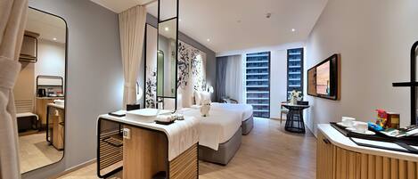A modern hotel room with a large bed, a desk with a mirror, a small table with a vase of flowers, and a view of the city through the window.