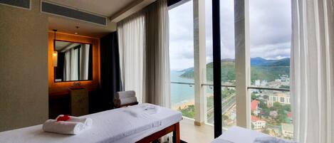Una habitación de hotel con dos camas, un ventanal con vistas a una ciudad costera y un espejo en la pared.