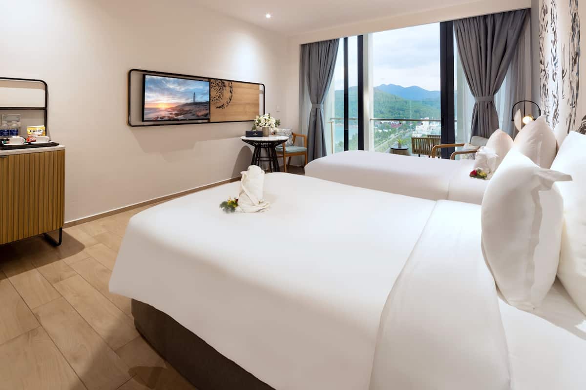 Ein Hotelzimmer mit zwei Betten, einem Schreibtisch mit Stuhl, einem kleinen Tisch ebenfalls mit Stuhl und einem großen Fenster mit Blick auf Berge.