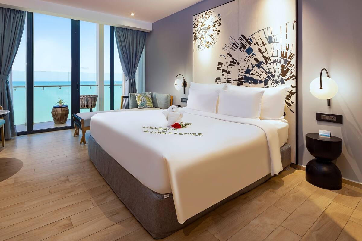Ein Hotelzimmer mit einem großen Bett, einem Balkon mit Meerblick, einem kleinen Tisch mit Stuhl und einer Wanddekoration.