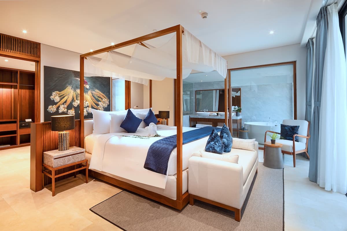 Ein geräumiges Schlafzimmer mit einem Himmelbett, einer Sitzecke inklusive Sofa und Sesseln, einem großen Spiegel und Blick ins Bad.