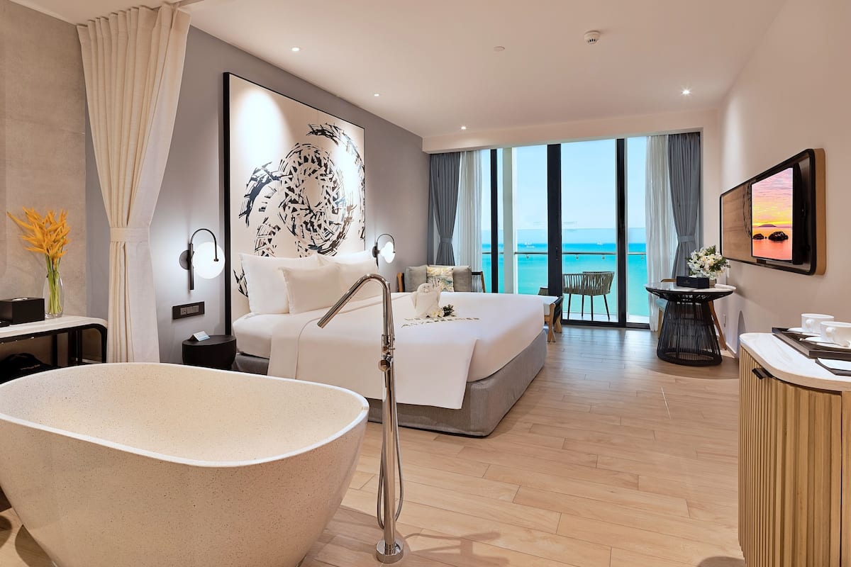 Ein modernes Hotelzimmer mit einer großen Badewanne, einem Bett mit weißen Laken, Meerblick durch große Fenster und einem Balkon mit Sitzgelegenheit.