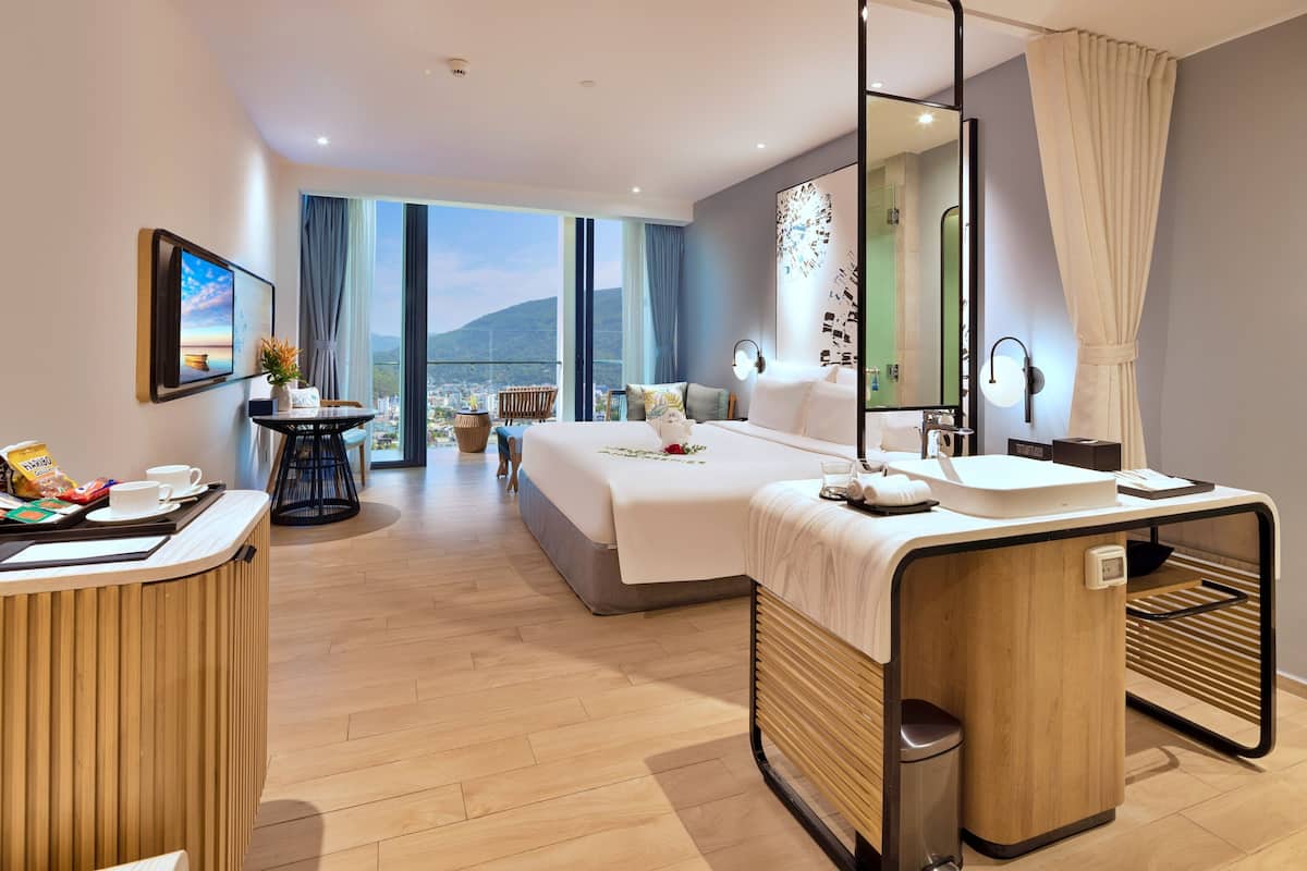 Ein modernes Hotelzimmer mit einem großen Bett, einem Schreibtisch mit Spiegel, einem kleinen Tisch mit Vase und Pflanze sowie Blick auf Stadt und Berge durch große Fenster.