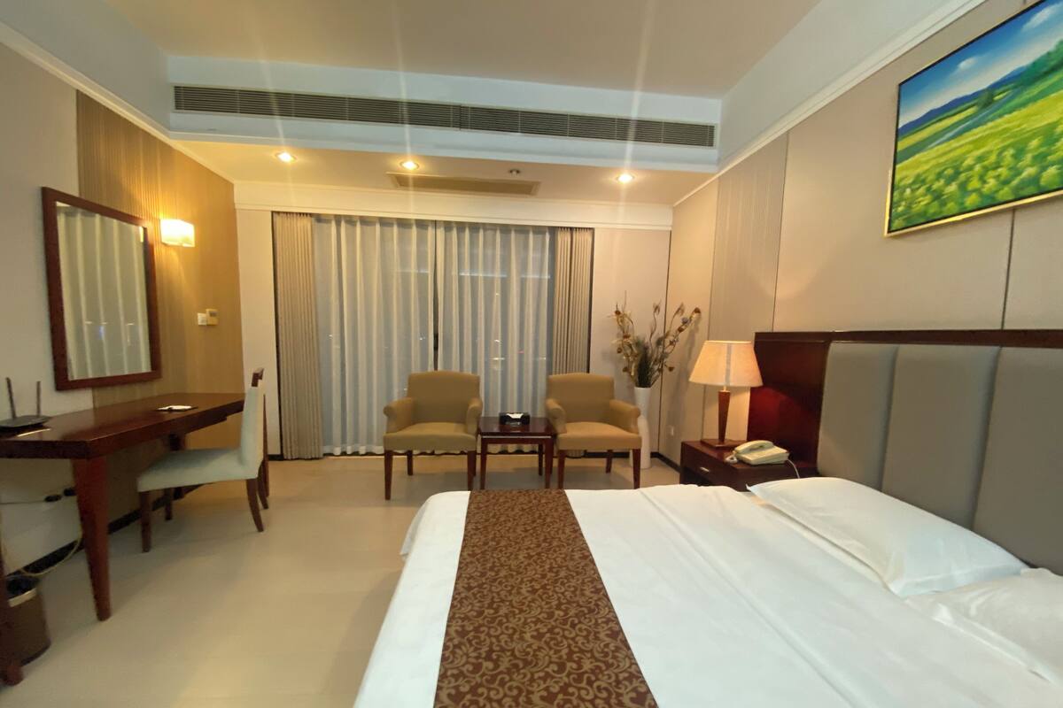 Habitación de hotel con cama, dos sillones, escritorio, lámpara, teléfono y un cuadro en la pared.