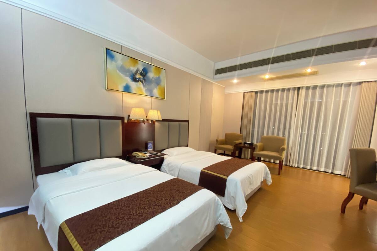 Habitación de hotel con dos camas, un escritorio, dos sillas, un cuadro y una ventana con cortinas.