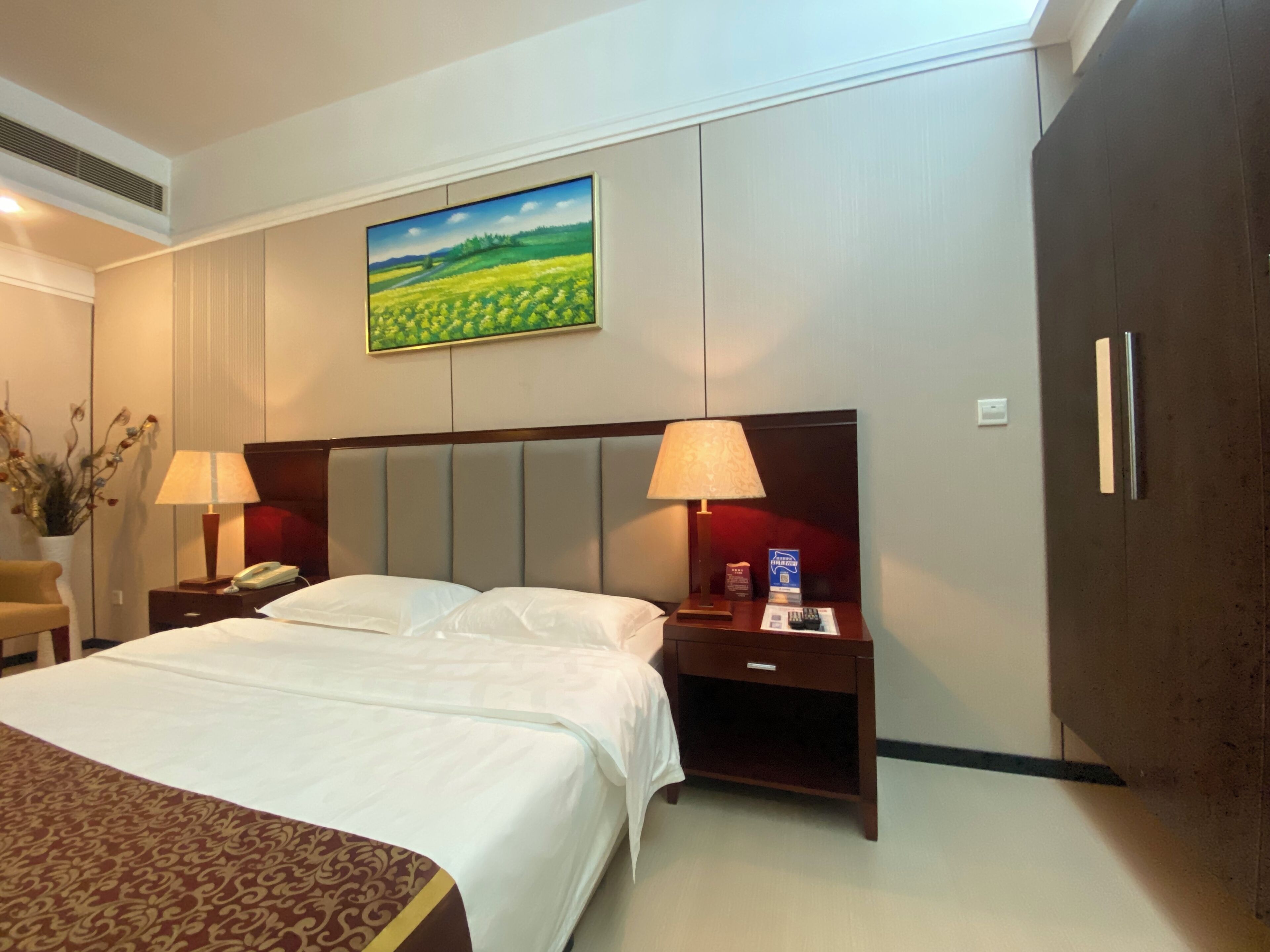 A hotel room with a large bed, two bedside tables with lamps, a nightstand with a phone and a remote control, a wardrobe, and a painting on the wall.