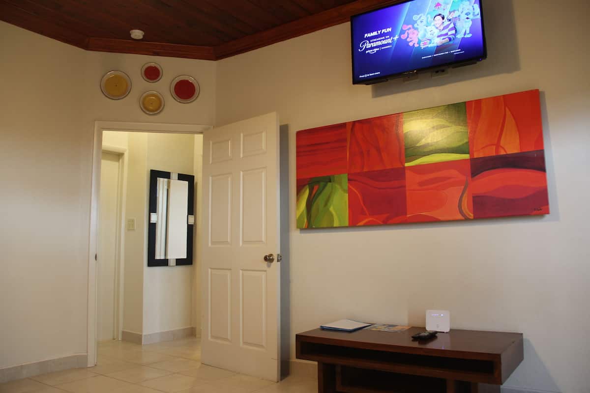 Um corredor com uma televisão presa à parede exibindo um programa, uma pintura abstrata colorida e uma mesinha com um controle remoto.