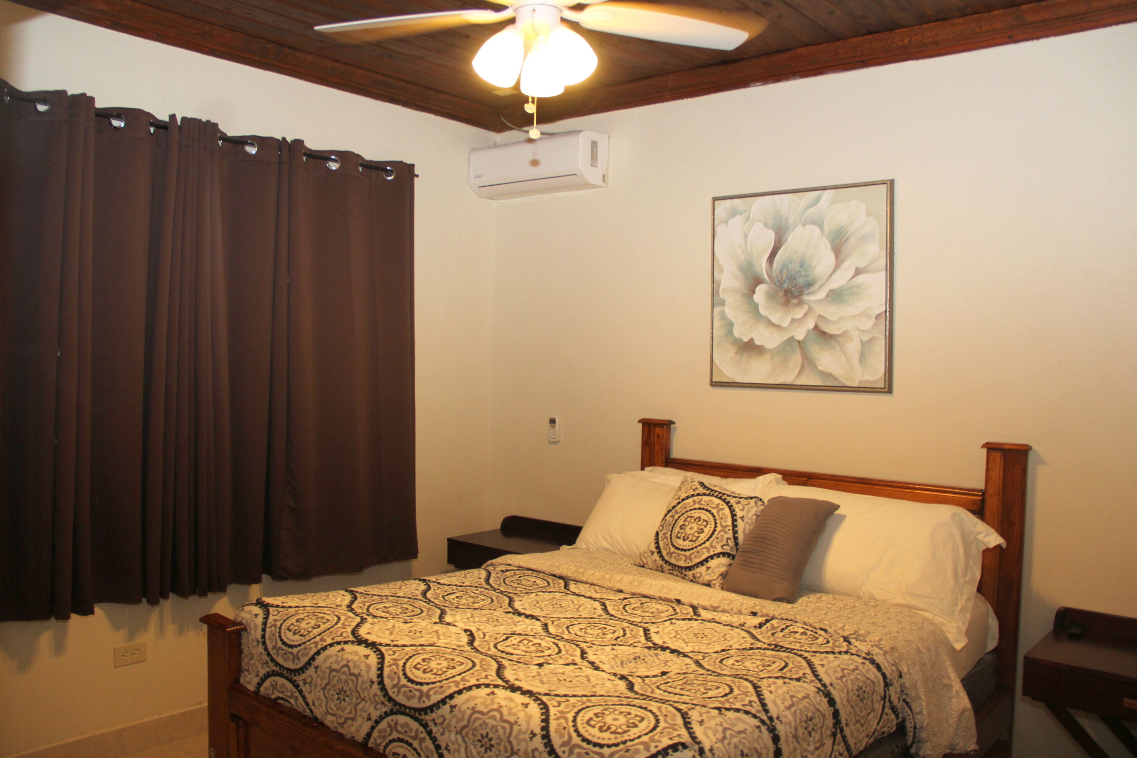 Un dormitorio con cama, ventilador de techo, equipo de aire acondicionado, mesita de noche y una pared con un cuadro floral.