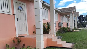 Un edificio de dos plantas con fachada rosa y detalles blancos. Hay escalones que llevan a una puerta con ventanal, y un pequeño jardín con plantas y un sendero.