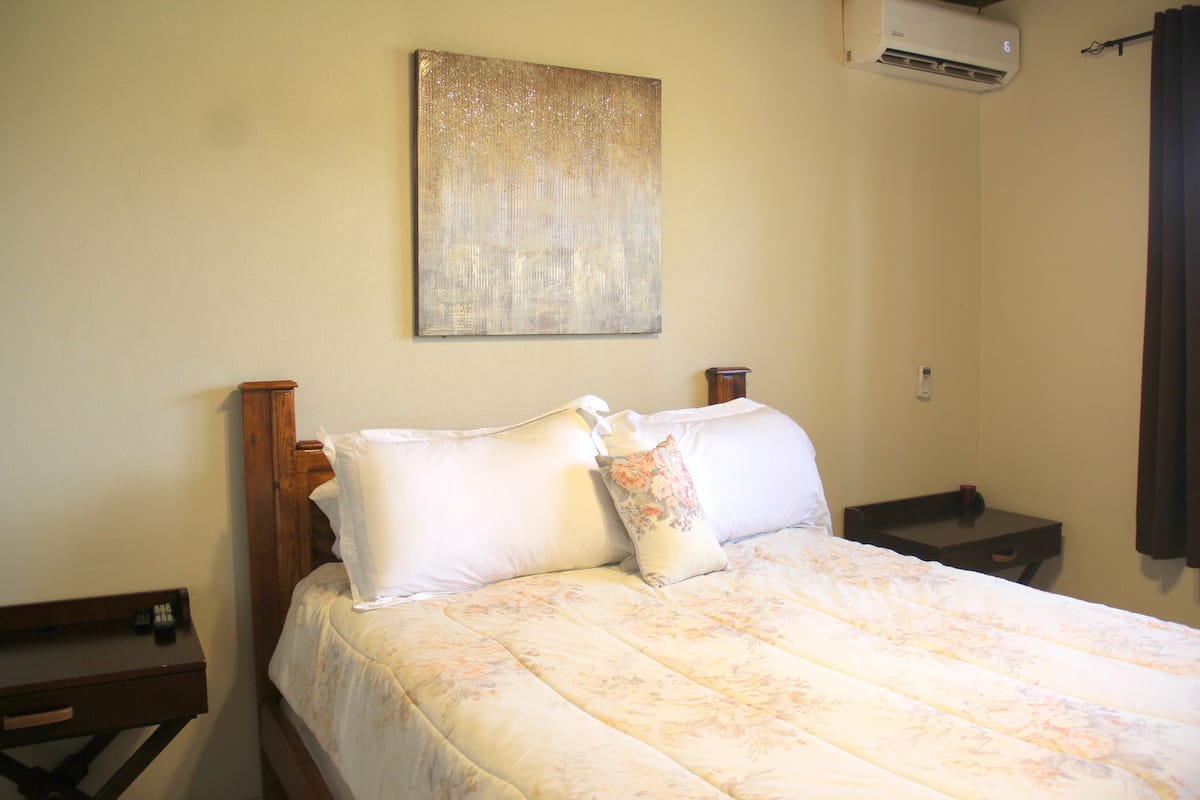Uma cama bem arrumada com edredom floral e duas almofadas, uma pequena mesa de cabeceira com abajur e um ar condicionado fixado na parede.