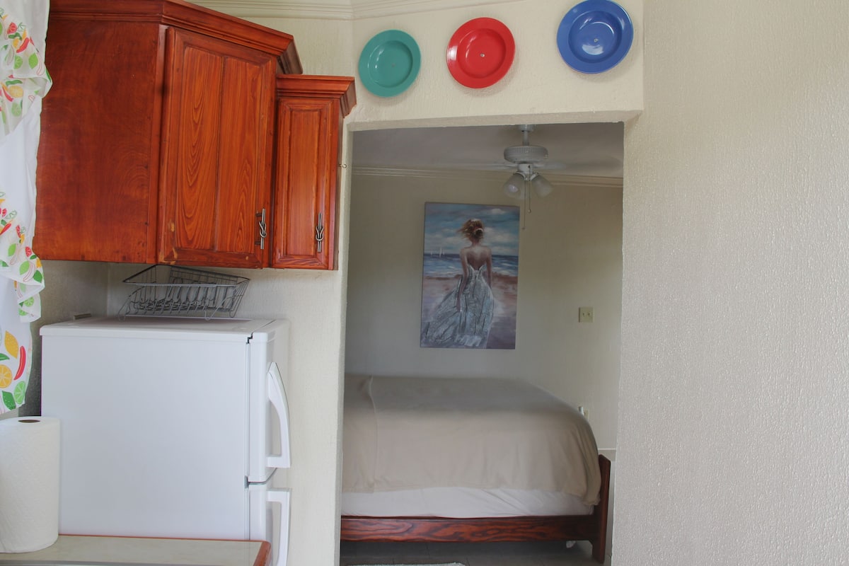 Quarto compacto com uma cama, um armário de madeira e um quadro com uma paisagem praiana.