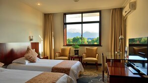 A hotel room with two beds, a desk, a TV, and a view of mountains and greenery. - Jiuhuashan Center Hotel (Chizhou)