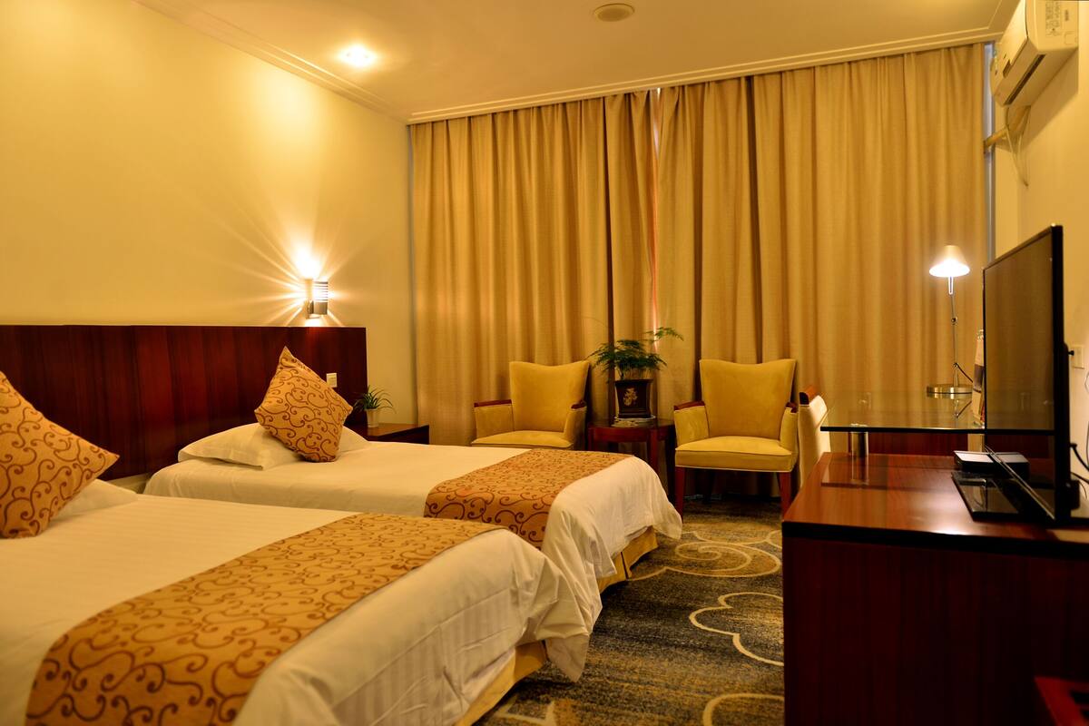 A hotel room with two beds, a desk, a chair, a television, and a lamp.