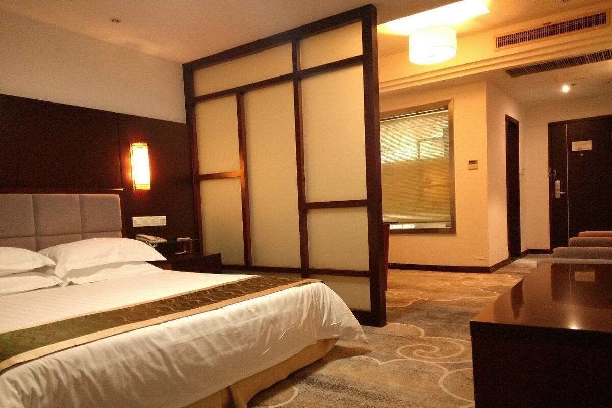 A hotel room with a large bed, a bedside table, a desk, and a window with blinds.